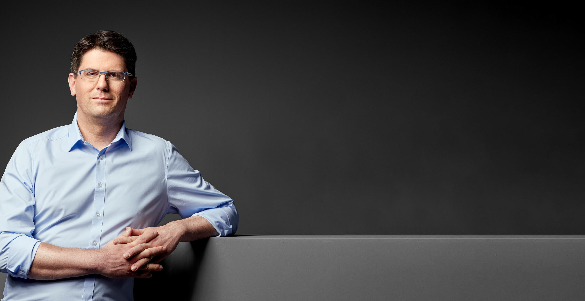 Foto: Prof. Christoph Hell lehnt an einem dunkelgrauen Mauervorsprung vor dunkelgrauem Hintergrund, Blick nach vorne 